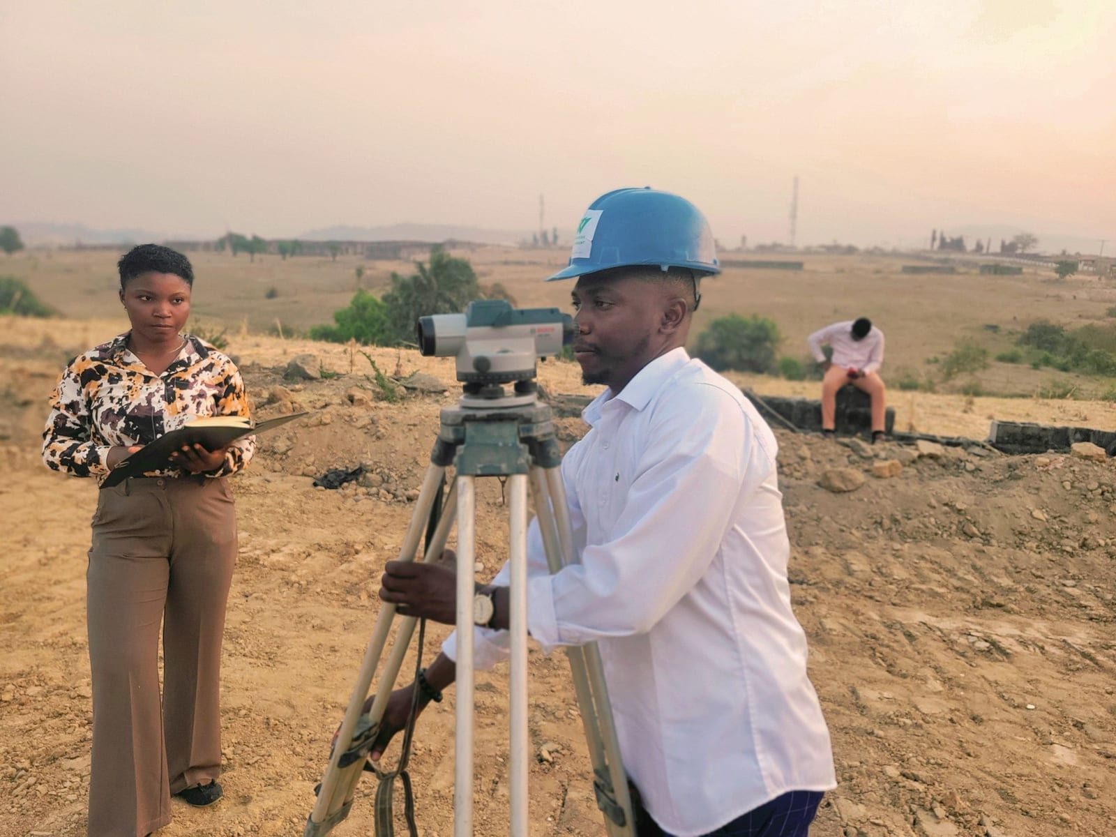 A man in a blue hard hat looks through a surveyor's level on cleared land while a woman in a patterned blouse holds a notebook.