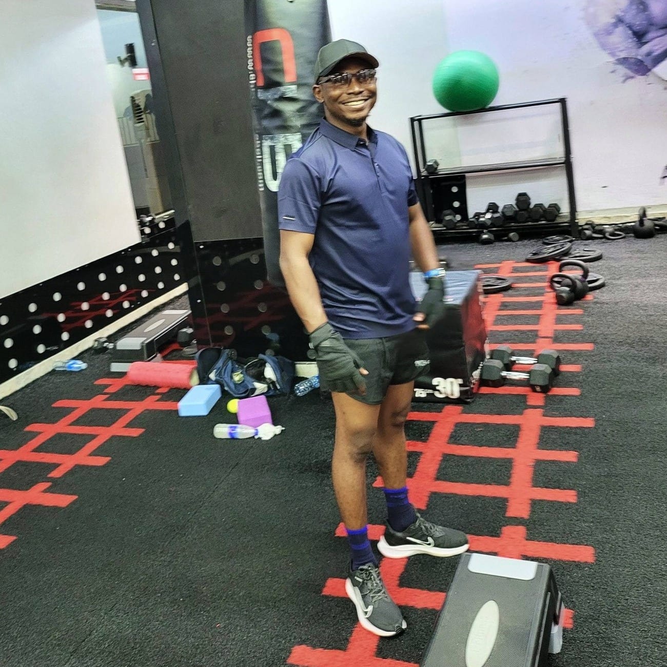 A smiling man in a cap, glasses, navy polo shirt, shorts and training gloves stands in a gym among dumbbells, kettlebells and a punching bag.