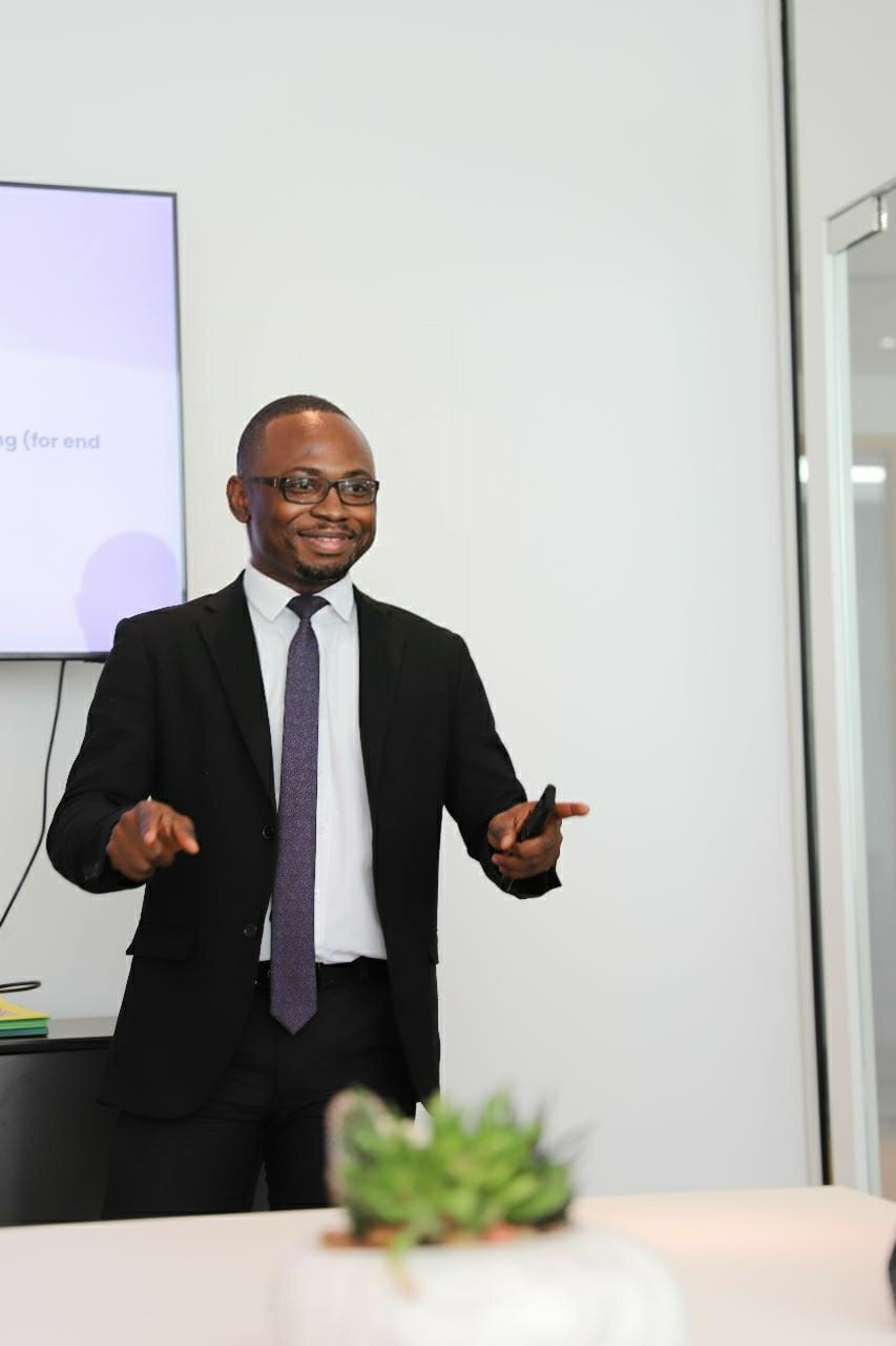 A smiling man in glasses, a dark suit and purple tie gestures as he presents, clicker in hand, beside a wall-mounted screen.