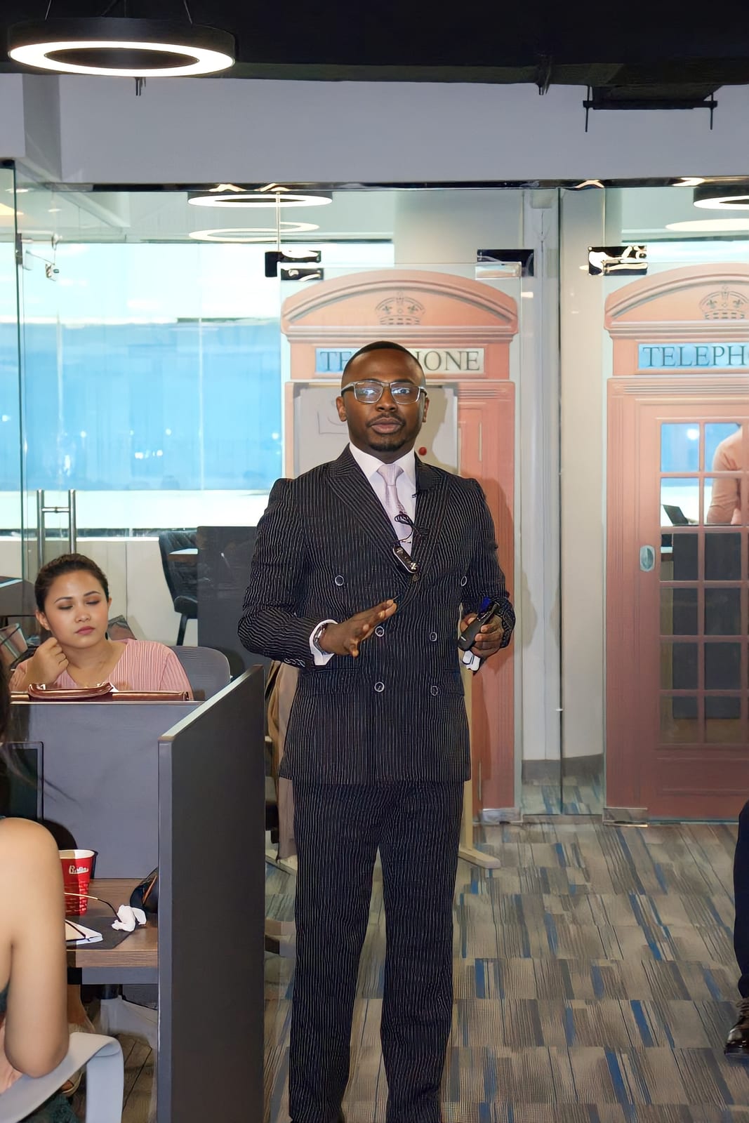 A man in glasses and a pinstripe double-breasted suit speaks to people at desks in an office with red telephone-box booths.