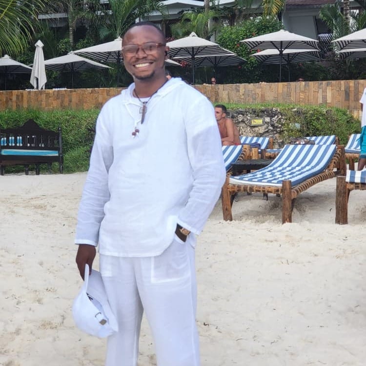 A smiling man in white linen and glasses, holding a white cap, stands on a sandy beach with striped sun loungers and parasols.