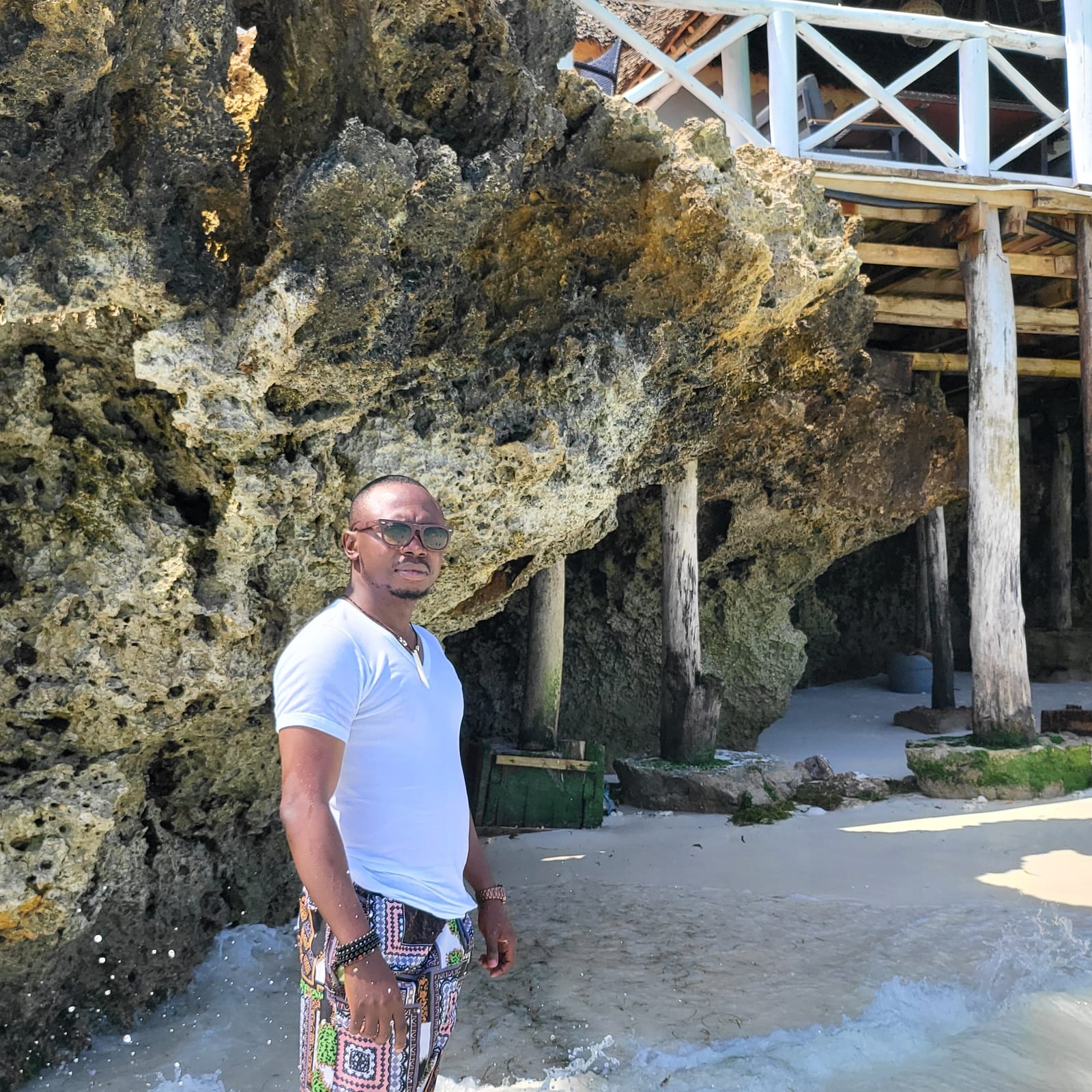 A man in sunglasses, a white T-shirt and patterned trousers stands in shallow surf beside a coral rock outcrop under a wooden deck on stilts.