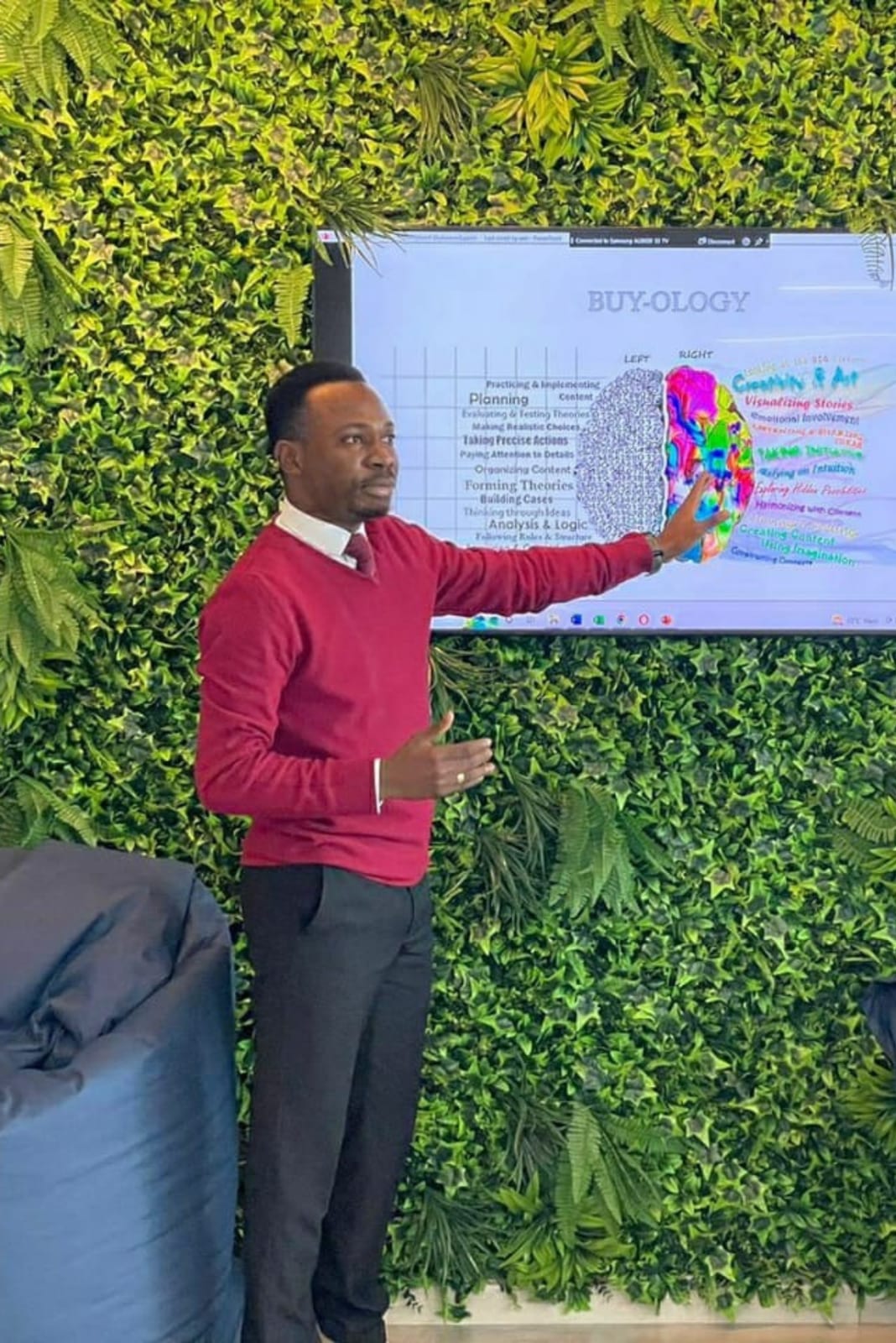 A man in a red sweater and tie points to a 'Buy-ology' left-brain, right-brain slide on a screen set in a green plant wall.