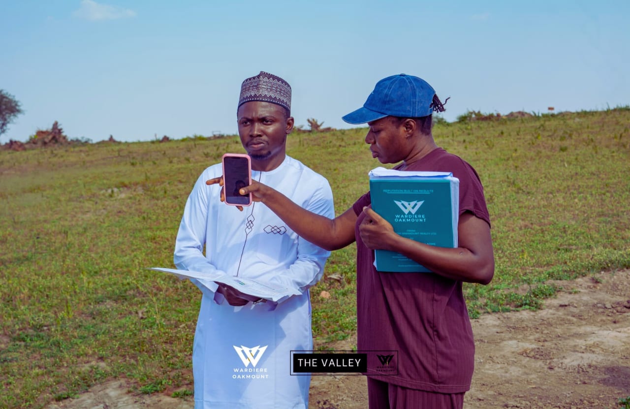 A woman holding a Wardiere Oakmount folder shows her phone to a man in a white kaftan with a printed plan, on open land; captioned 'The Valley'.