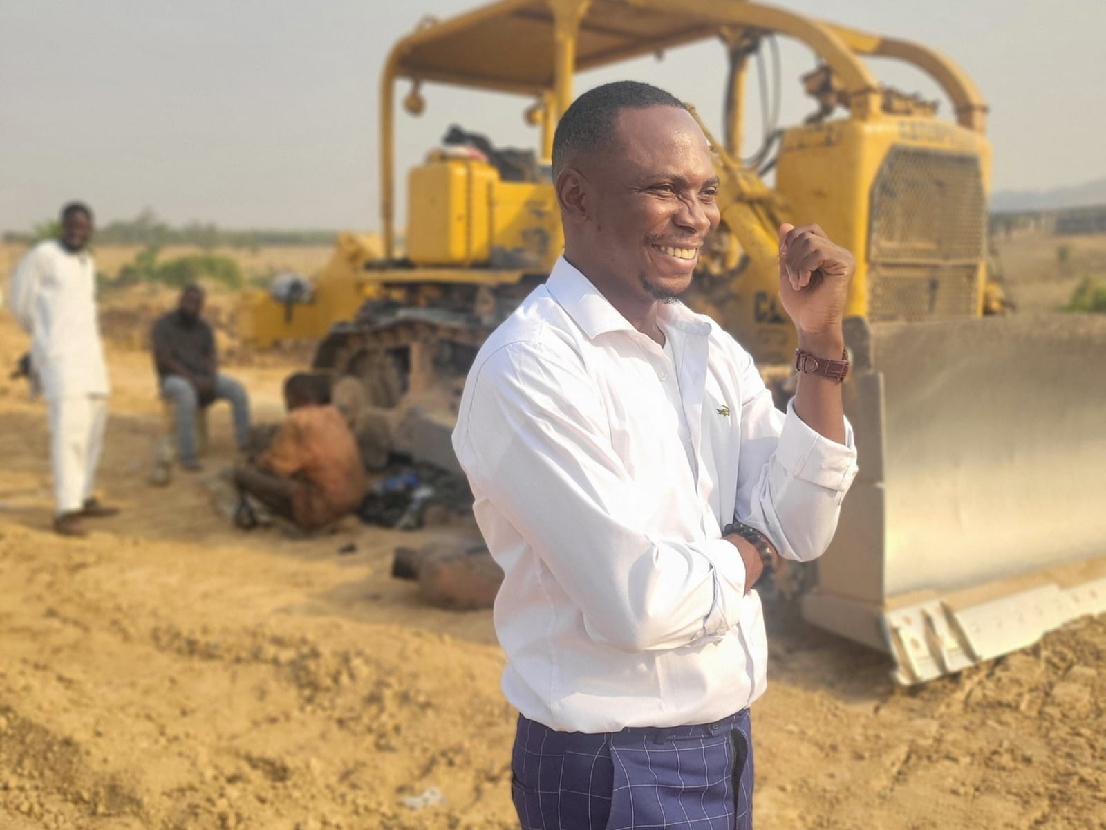 A smiling man in a white shirt stands in front of a yellow bulldozer on cleared land, with workers nearby.