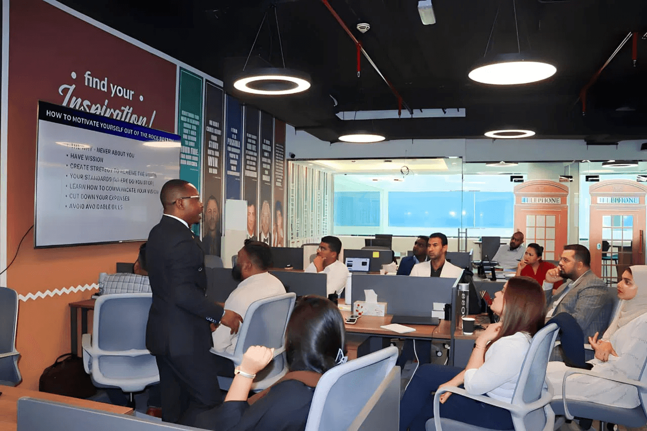 A man in a dark suit presenting to seated staff in a Dubai real estate office, the screen reading “How to motivate yourself out of the rock bottom”.