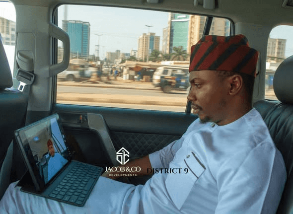 A man in a striped cap and white agbada takes a founder's video call on a tablet in a car, watermarked Jacob & Co Developments District 9.